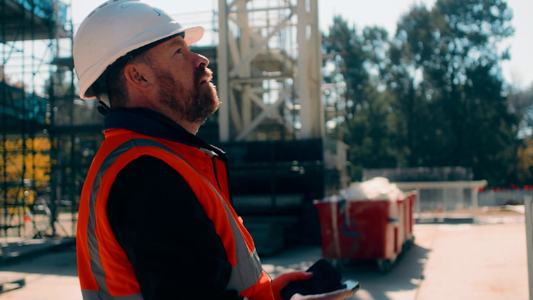 a contractor in a hard hat and vest holding a tablet while looking up at the construction site